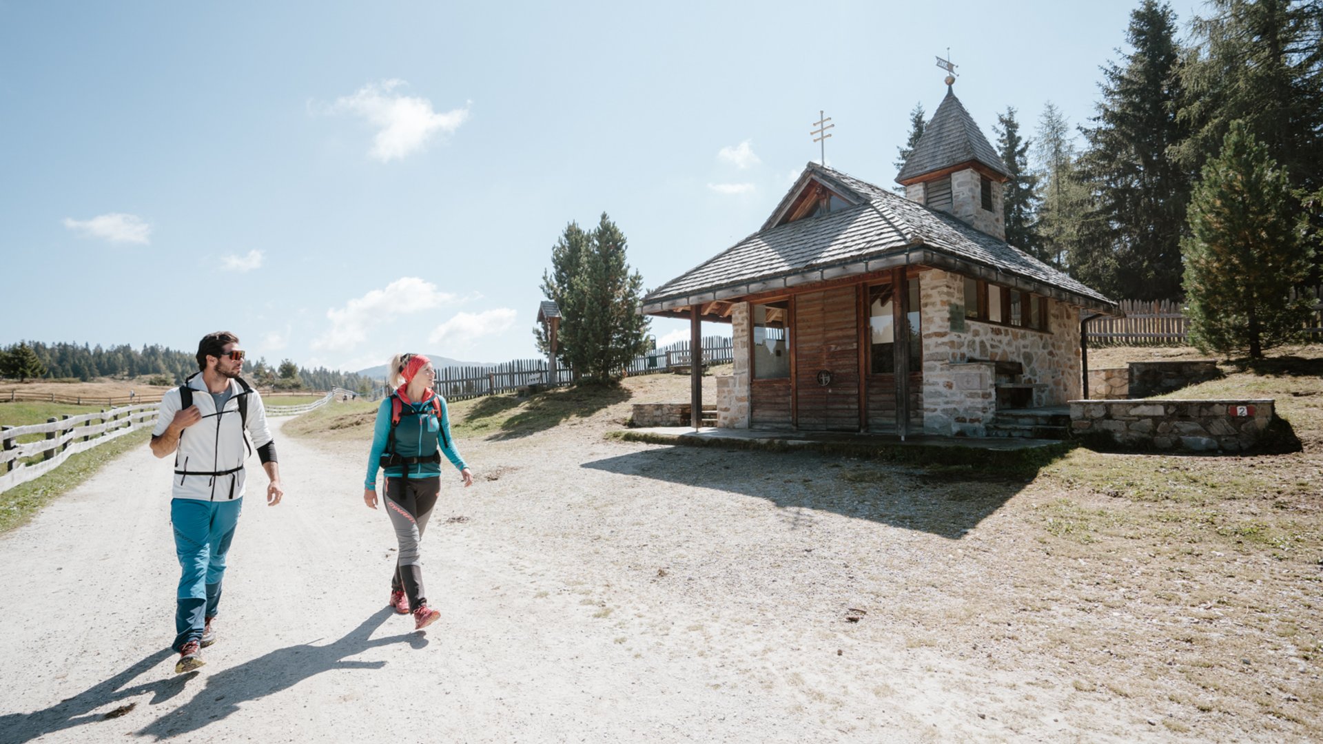 Trekking sulle Dolomiti L'immagine mostra due escursionisti, un uomo e una donna, che camminano lungo un ampio sentiero sabbioso. Alla loro destra c'è una piccola cappella in pietra con il tetto in legno. Il cielo è limpido e azzurro, con dolci colline e alberi sullo sfondo.